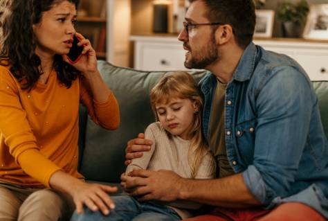 Sad little girl sitting between parents on sofa while her mother talking on phone with social worker about custody and child support after divorce
